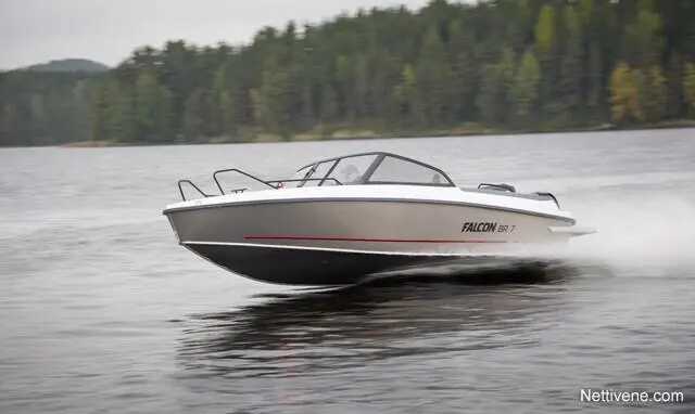 A sleek Falcon boat speeds across a lake, creating ripples, surrounded by lush green trees and cloudy skies.